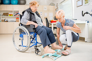 A female nursing home patient in a wheelchair being assisted with foot exercises by a female healthcare worker